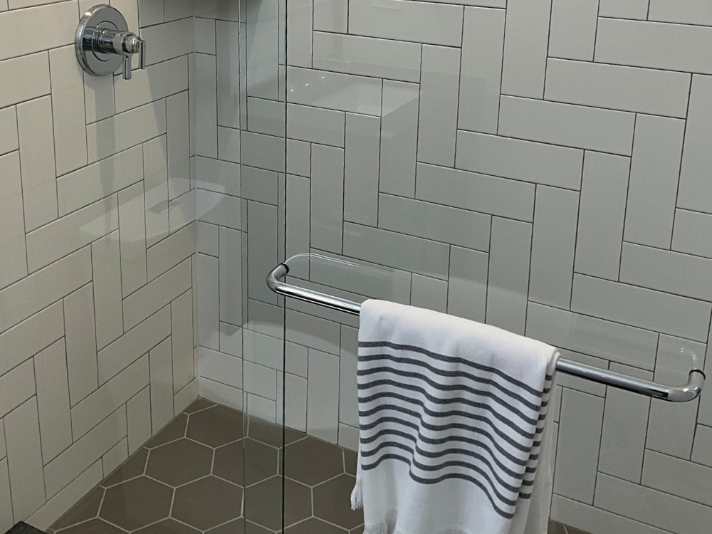 A shower stall with white subway tile in a herringbone pattern on the walls and hexagonal gray tiles on the floor. A glass door has a silver handle with a striped towel hanging from it.